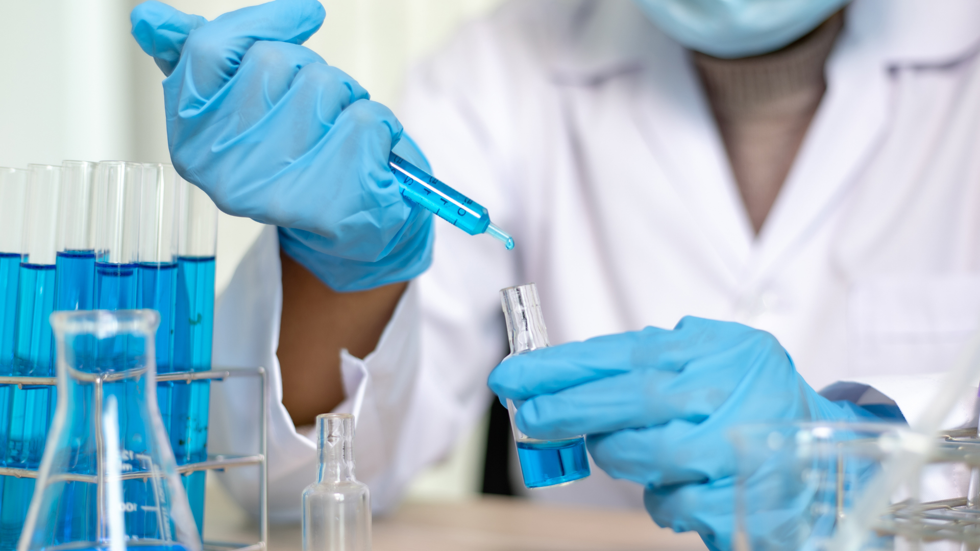 Associate scientist performing lab work in formulation development, pipetting blue solution into a glass vial with test tubes in the background.