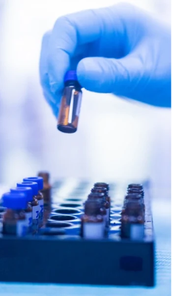 Gloved hand holding a small vial above a tray of sample vials in a laboratory, representing analytical work in drug formulation at SeraNovo.